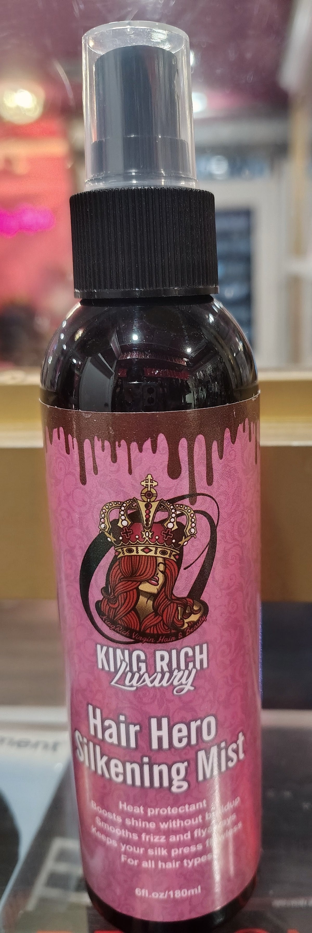 A bottle of King Rich Luxury Hair Hero Silkening Mist with a pink label and black spray top, shown against a blurred indoor background.